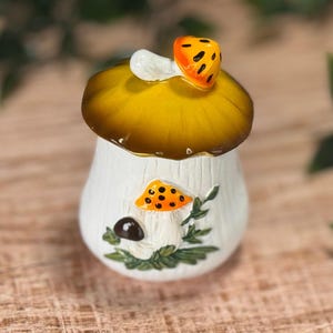 Puede incluir: Un tarro de cer&aacute;mica blanca con una tapa en forma de sombrero de seta grande de color amarillo oscuro. El tarro presenta un relieve en la parte frontal que muestra una seta naranja m&aacute;s peque&ntilde;a con puntos negros, una seta marr&oacute;n y hojas verdes.