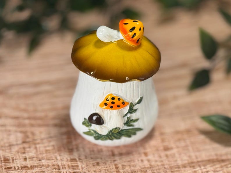 Puede incluir: Un tarro de cer&aacute;mica blanca con una tapa en forma de sombrero de seta grande de color amarillo oscuro. El tarro presenta un relieve en la parte frontal que muestra una seta naranja m&aacute;s peque&ntilde;a con puntos negros, una seta marr&oacute;n y hojas verdes.