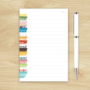 May include: A white notepad with a lined page and a white pen. The notepad has a decorative border with colorful bowls with different patterns.