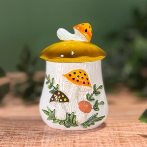 Puede incluir: Un tarro de cer&aacute;mica blanca con una tapa en forma de seta amarilla. El cuerpo del tarro presenta ilustraciones en relieve de varias setas en naranja, marr&oacute;n y beige, acentuadas con hojas y enredaderas verdes con un dise&ntilde;o bot&aacute;nico.