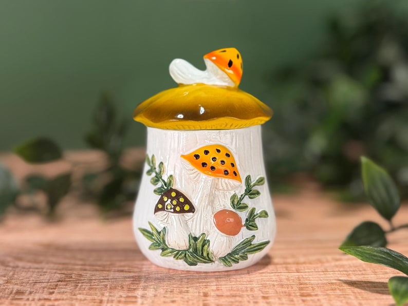 Puede incluir: Un tarro de cer&aacute;mica blanca con una tapa en forma de seta amarilla. El cuerpo del tarro presenta ilustraciones en relieve de varias setas en naranja, marr&oacute;n y beige, acentuadas con hojas y enredaderas verdes con un dise&ntilde;o bot&aacute;nico.