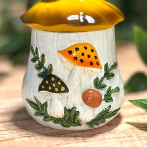 Puede incluir: Un tarro de cer&aacute;mica con una tapa en forma de sombrero de seta amarillo. La base blanca presenta un dise&ntilde;o en relieve pintado a mano de tres setas en naranja, marr&oacute;n y verde, rodeadas de enredaderas y follaje verde sobre una superficie.