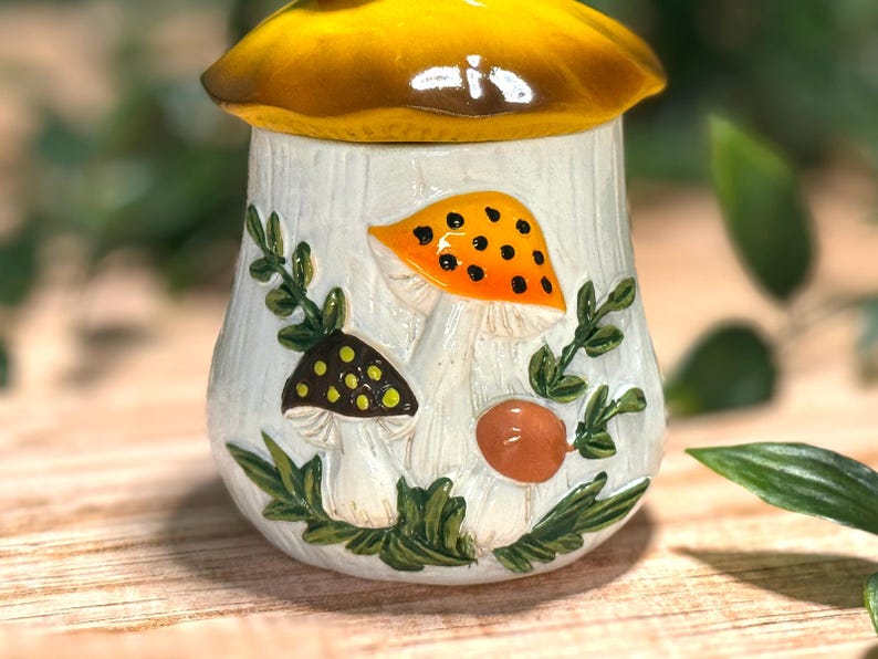 Puede incluir: Un tarro de cer&aacute;mica con una tapa en forma de sombrero de seta amarillo. La base blanca presenta un dise&ntilde;o en relieve pintado a mano de tres setas en naranja, marr&oacute;n y verde, rodeadas de enredaderas y follaje verde sobre una superficie.