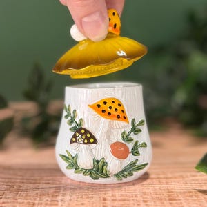 Puede incluir: Un tarro de cer&aacute;mica blanca con una tapa extra&iacute;ble en forma de sombrero de seta amarilla. El tarro presenta ilustraciones en relieve de varias setas en naranja, marr&oacute;n y amarillo, acentuadas con hojas y enredaderas verdes en la base.