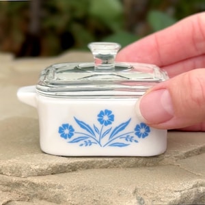 May include: A small rectangular white glass butter dish with a clear glass lid and a knob handle. The front of the dish features a blue floral pattern consisting of three stylized flowers with leaves and stems centered on the white surface.