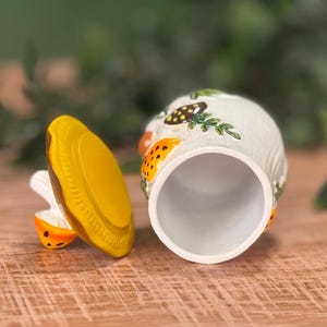 Puede incluir: Un tarro de cer&aacute;mica con tapa extra&iacute;ble, que presenta una base blanca decorada con setas pintadas en naranja y marr&oacute;n y follaje verde. La tapa es de color amarillo s&oacute;lido con un borde texturizado y el tarro tiene un interior cil&iacute;ndrico hueco.