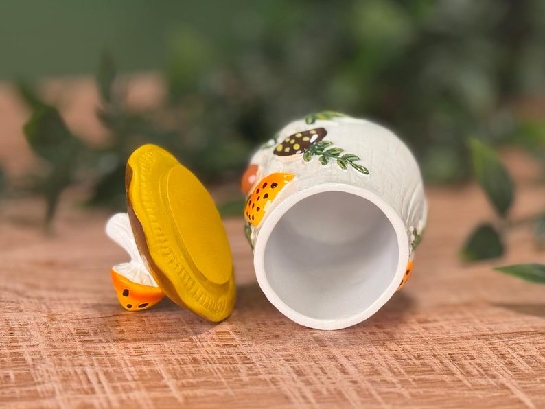 Puede incluir: Un tarro de cer&aacute;mica con tapa extra&iacute;ble, que presenta una base blanca decorada con setas pintadas en naranja y marr&oacute;n y follaje verde. La tapa es de color amarillo s&oacute;lido con un borde texturizado y el tarro tiene un interior cil&iacute;ndrico hueco.