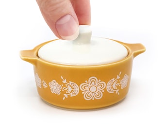 Puede incluir: Una pequeña cazuela de cerámica redonda de color amarillo mostaza con dos asas laterales y una tapa blanca. El exterior del recipiente presenta un patrón floral blanco repetitivo que rodea la base. La tapa tiene un pequeño pomo central.