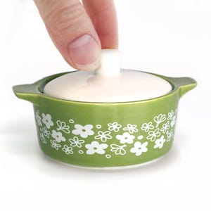 May include: A small, green ceramic casserole dish with a white lid. The dish is decorated with white floral patterns. The lid has a white knob. The dish has two small handles on either side.