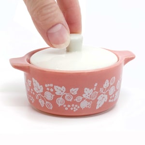 May include: A small, pink ceramic casserole dish with a white lid. The dish features a white floral pattern around the exterior. The lid is being lifted by a hand. The dish has two small handles on either side.
