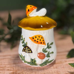 Puede incluir: Un tarro de cer&aacute;mica con una tapa en forma de gran sombrero de seta amarillo-marr&oacute;n coronado por una seta naranja m&aacute;s peque&ntilde;a. El cuerpo cil&iacute;ndrico blanco presenta ilustraciones en relieve de setas naranjas, marrones y blancas.