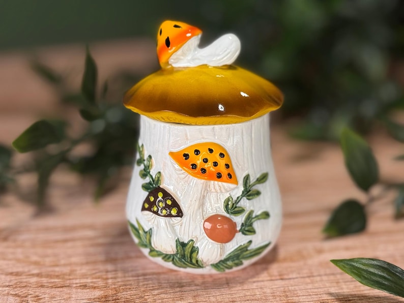Puede incluir: Un tarro de cer&aacute;mica con una tapa en forma de gran sombrero de seta amarillo-marr&oacute;n coronado por una seta naranja m&aacute;s peque&ntilde;a. El cuerpo cil&iacute;ndrico blanco presenta ilustraciones en relieve de setas naranjas, marrones y blancas.