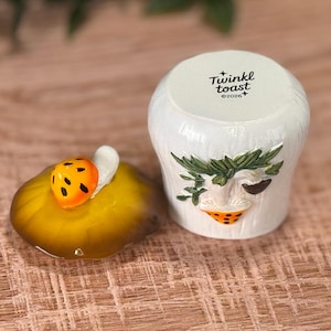 Puede incluir: Un tarro de cer&aacute;mica blanca con una tapa que presenta una peque&ntilde;a mariquita naranja. El cuerpo del tarro tiene una superficie texturizada con un dise&ntilde;o esculpido de una hoja verde y una seta naranja. La tapa tiene impreso Twinkl toast y 2026.