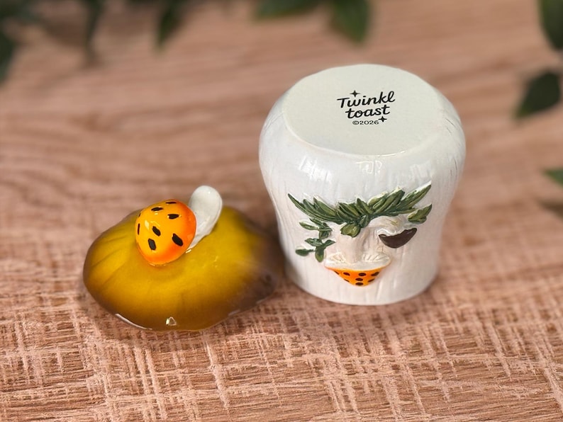 Puede incluir: Un tarro de cer&aacute;mica blanca con una tapa que presenta una peque&ntilde;a mariquita naranja. El cuerpo del tarro tiene una superficie texturizada con un dise&ntilde;o esculpido de una hoja verde y una seta naranja. La tapa tiene impreso Twinkl toast y 2026.