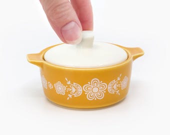 May include: A small, round, mustard-yellow ceramic casserole dish with a white lid. The dish has a white floral pattern and two small handles. The lid is being lifted by a hand.