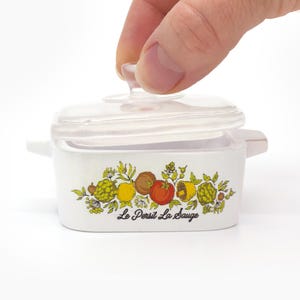 May include: A small, white casserole dish with a clear lid. The dish is decorated with a colorful fruit and vegetable design and the text "Le Persil La Sauge". The lid is being lifted by a hand.