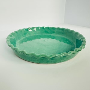 May include: A round, shallow ceramic pie dish with a vibrant, glossy turquoise green glaze. The dish features a distinctively wavy, scalloped edge along the entire rim, creating a decorative border around the smooth, circular base of the vessel.
