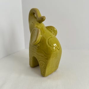 K&ouml;nnte beinhalten: Eine chartreusefarbene Elefantenfigur aus Keramik mit gl&auml;nzender Craquel&eacute;-Glasur. Der Elefant ist stehend dargestellt, wobei der R&uuml;ssel nach oben gerichtet ist. Die Oberfl&auml;che weist dezente, gepr&auml;gte Kreis- und Wellenlinienmuster auf.