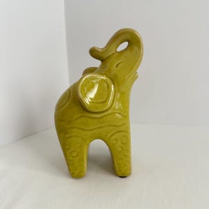 K&ouml;nnte beinhalten: Eine chartreusefarbene Elefantenfigur aus Keramik mit einer gl&auml;nzenden, rissigen Glasur. Der Elefant ist stehend mit erhobenem R&uuml;ssel dargestellt und weist gepr&auml;gte kreisf&ouml;rmige sowie wellenf&ouml;rmige Muster auf seinem K&ouml;rper und seinen Ohren auf.