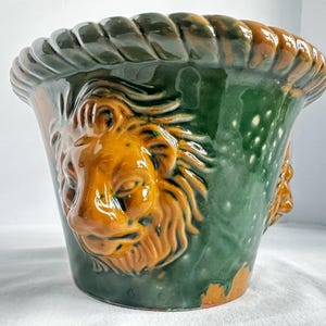 May include: A glazed ceramic planter featuring a deep green body with a prominent, raised, amber-colored lion head relief on the side. The rim of the vessel is designed with a thick, twisted rope texture in alternating shades of green and amber.