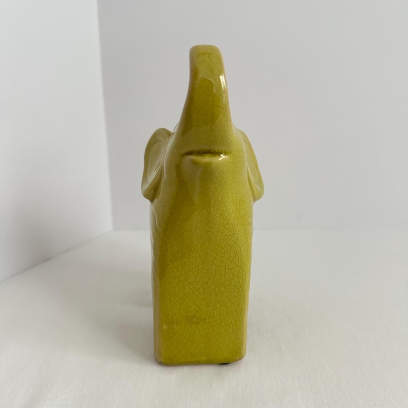 K&ouml;nnte beinhalten: Eine chartreusefarbene Elefantenfigur aus Keramik mit einer gl&auml;nzenden, rissigen Glasuroberfl&auml;che. Der Elefant ist in stehender Position mit erhobenem R&uuml;ssel dargestellt und zeigt eine glatte, abgerundete Form mit sichtbaren feinen Linien.