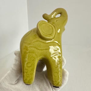 K&ouml;nnte beinhalten: Eine Elefantenfigur aus Keramik mit einer gl&auml;nzenden, chartreuse-gr&uuml;nen Glasur und einer sichtbaren Craquel&eacute;-Oberfl&auml;che. Der Elefant ist in stehender Position dargestellt, mit dem R&uuml;ssel nach oben gebogen und den Ohren zur Seite gerichtet.