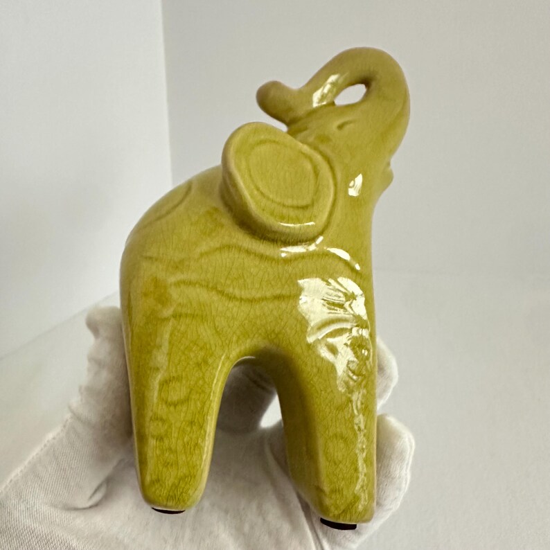 K&ouml;nnte beinhalten: Eine Elefantenfigur aus Keramik mit einer gl&auml;nzenden, chartreuse-gr&uuml;nen Glasur und einer sichtbaren Craquel&eacute;-Oberfl&auml;che. Der Elefant ist in stehender Position dargestellt, mit dem R&uuml;ssel nach oben gebogen und den Ohren zur Seite gerichtet.