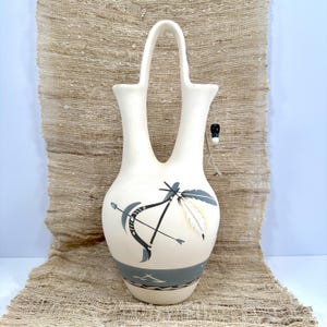 May include: A cream-colored ceramic wedding vase with a double-spout design and a central arched handle. The body features a painted black bow and arrow graphic, a white feather accent, and a gray band at the base with thin black line details.