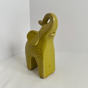 K&ouml;nnte beinhalten: Eine chartreusefarbene Elefantenfigur aus Keramik mit einer gl&auml;nzenden, rissigen Glasur. Der Elefant ist in stehender Position mit erhobenem R&uuml;ssel dargestellt. Der K&ouml;rper weist dezente, gepr&auml;gte wellenf&ouml;rmige Linienmuster an den Seiten auf.