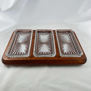 May include: A rectangular wooden serving board featuring three inset clear glass dishes. Each glass dish has a pressed sunburst pattern radiating from the center and a scalloped edge. The wood base has a smooth, dark brown finish.