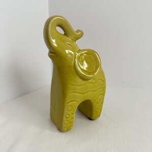 K&ouml;nnte beinhalten: Eine chartreusefarbene Keramik-Elefantenfigur mit gl&auml;nzender Craquel&eacute;-Glasur. Der Elefant ist stehend dargestellt, wobei der R&uuml;ssel nach oben gerichtet ist. Der K&ouml;rper weist gepr&auml;gte Wellenlinien und kreisf&ouml;rmige Muster an den Beinen auf.
