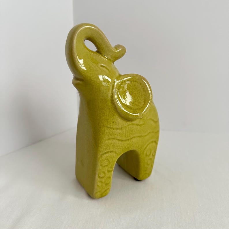 K&ouml;nnte beinhalten: Eine chartreusefarbene Keramik-Elefantenfigur mit gl&auml;nzender Craquel&eacute;-Glasur. Der Elefant ist stehend dargestellt, wobei der R&uuml;ssel nach oben gerichtet ist. Der K&ouml;rper weist gepr&auml;gte Wellenlinien und kreisf&ouml;rmige Muster an den Beinen auf.