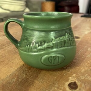 May include: A sage green ceramic mug featuring a rounded body and a single handle. The exterior displays a molded relief of a building surrounded by trees, with an oval emblem at the base containing the embossed letters GPI in a serif font.