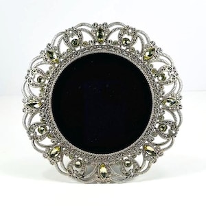 May include: A round picture frame with a metallic silver finish and an intricate filigree border. The frame features a central circular black velvet insert surrounded by small, light-colored, marquise-cut crystals embedded in the metal scrollwork.