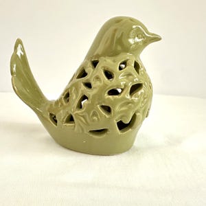May include: A sage green ceramic bird figurine featuring a glossy finish and intricate cut-out leaf patterns across its body. The bird is depicted in a seated position with its tail feathers pointed upward and its head turned to the right side.