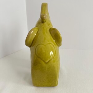 K&ouml;nnte beinhalten: Eine chartreusefarbene Elefantenfigur aus Keramik mit einer gl&auml;nzenden, rissigen Glasur. Der Elefant ist in stehender Position mit erhobenem R&uuml;ssel dargestellt. Eine markante Herzform ist in der Mitte seines K&ouml;rpers eingepr&auml;gt.
