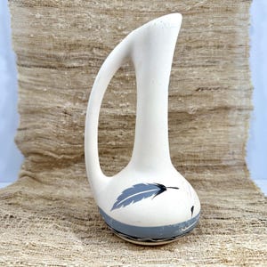 May include: A matte off-white ceramic vase with a tall, slender neck and a single arched handle. The rounded base features a painted grey feather design, a horizontal grey band, and thin black line detailing near the bottom edge of the vessel.