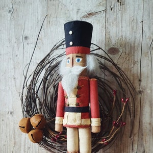 May include: A handmade felt nutcracker doll with a red and white uniform, black boots, and a white beard. The doll is hanging on a twig wreath with brown bells.