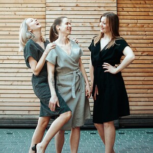 Op de afbeelding: Drie vrouwen in verschillende stijlen jurken, lachend en naar elkaar kijkend. De vrouwen staan voor een houten muur.