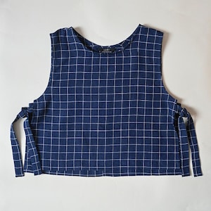 May include: A sleeveless, navy blue top with a white grid pattern. The top has tie closures on each side and the brand name "LUCI" is visible on the back of the neck. The fabric appears to be lightweight.