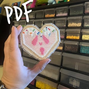 May include: A heart-shaped cross-stitch design with a pink center and a tan border. The design features pink shapes and colorful sprinkles. The word "PDF" is written in white script in the upper left corner. The background shows storage containers.