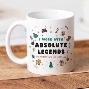Può includere: Tazza in ceramica bianca con la scritta "I WORK WITH ABSOLUTE LEGENDS" e la frase "(YES MY SECRET SANTA GAVE ME THIS MUG)". Decorata con illustrazioni natalizie colorate, tra cui un regalo, un biscotto e un albero di Natale.