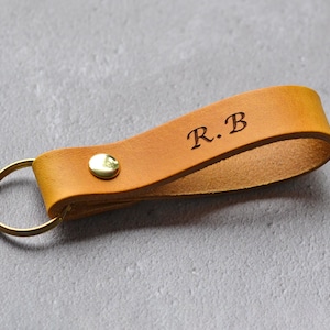 Könnte beinhalten: Ein beigefarbener Schlüsselanhänger aus Leder mit den eingeprägten Initialen R.B. Das Lederband ist mit einer glänzenden, goldfarbenen Metallniete befestigt und enthält einen passenden runden, goldfarbenen Metallring für Schlüssel.