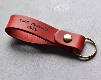 Puede incluir: Un llavero de cuero rojo con un botón de presión de metal dorado y una anilla circular de metal dorado. La correa de cuero tiene grabado el texto negro "YOUR MESSAGE HERE" en la superficie frontal del cuero doblado.