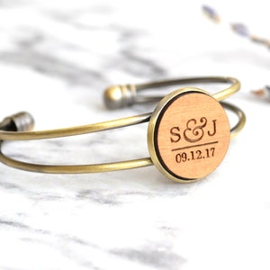Könnte beinhalten: Ein goldfarbenes Metall-Armreif-Armband mit einem runden Holzanhänger. Der Anhänger ist mit den Initialen "S&J" und dem Datum "09.12.17" graviert.
