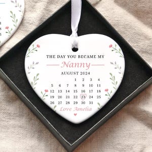 Op de afbeelding: Witte hartvormige keramische hanger met een bloemenontwerp en de tekst "The day you became my Nanny - August 2024" met een kalenderrooster dat de maand augustus laat zien. De hanger bevat ook de tekst "Love Amelia".
