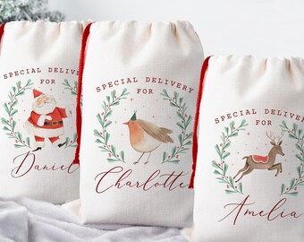 May include: Three off-white drawstring gift bags with red drawstrings. Each bag features a watercolor illustration within a green wreath and the words "Special Delivery For" above a name. One bag has Santa, another a bird, and the last a reindeer.