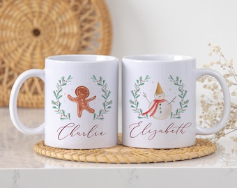 Könnte beinhalten: Zwei weiße Keramikbecher mit weihnachtlichen Motiven. Ein Becher zeigt einen Lebkuchenmann, der andere einen Schneemann, beide in einem grünen Kranz. Die Namen "Charlie" und "Elizabeth" sind in Schreibschrift geschrieben. Die Becher stehen auf einem gewebten Untersetzer.