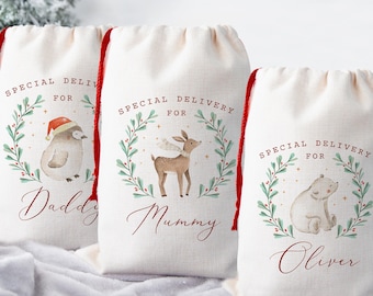 May include: Three off-white drawstring gift bags with red cords. Each bag features a watercolor illustration of a penguin, deer, and polar bear, respectively, surrounded by a green wreath. Text reads "Special Delivery For" and a name.