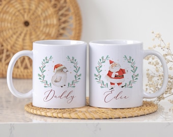 Könnte beinhalten: Zwei weiße Keramikbecher mit Weihnachtsdesigns. Ein Becher zeigt einen Pinguin mit Weihnachtsmütze und dem Wort "Daddy". Der andere Becher zeigt den Weihnachtsmann und das Wort "Edie". Beide haben einen grünen Kranz.